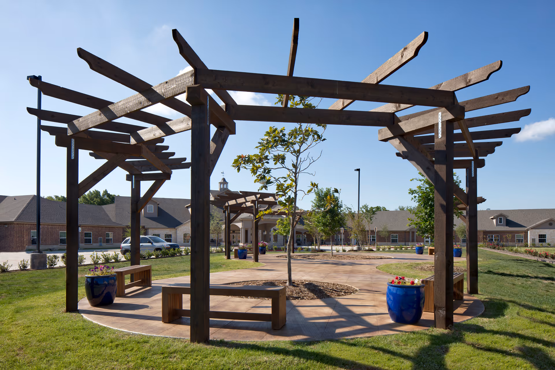 Outdoor seating area with wooden pergolas and benches surrounding small trees and blue flower pots, set on a paved circular patio with grass and a senior living facility building in the background under a clear blue sky.