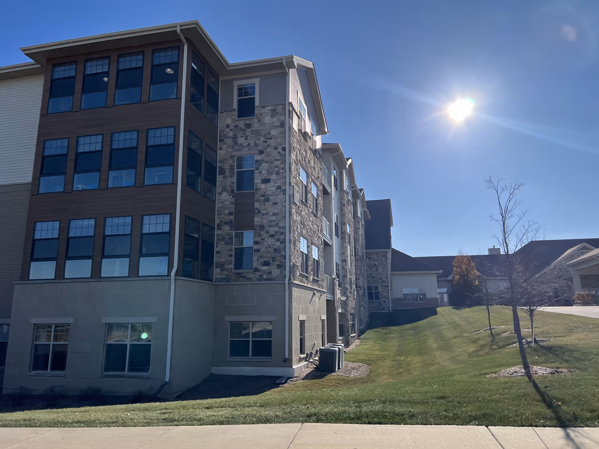 Exterior view of a multi-story senior living facility building with a mix of stone and wood siding under a clear blue sky with the sun shining brightly. There is a well-maintained grassy area with a few small trees in front of the building.