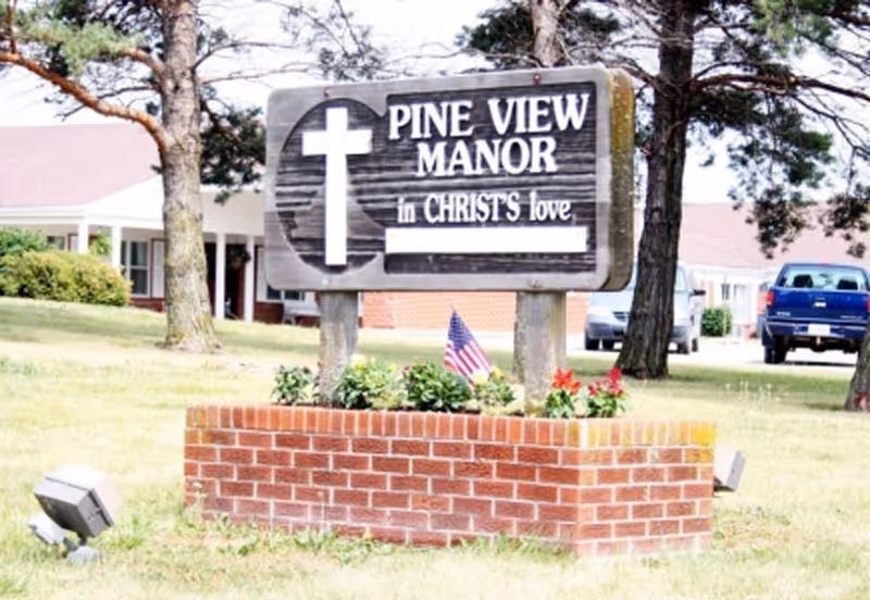 A wooden sign reading 'Pine View Manor in Christ's love' mounted above a brick planter with flowers and a small American flag on the lawn in front of a low building and parked vehicles.