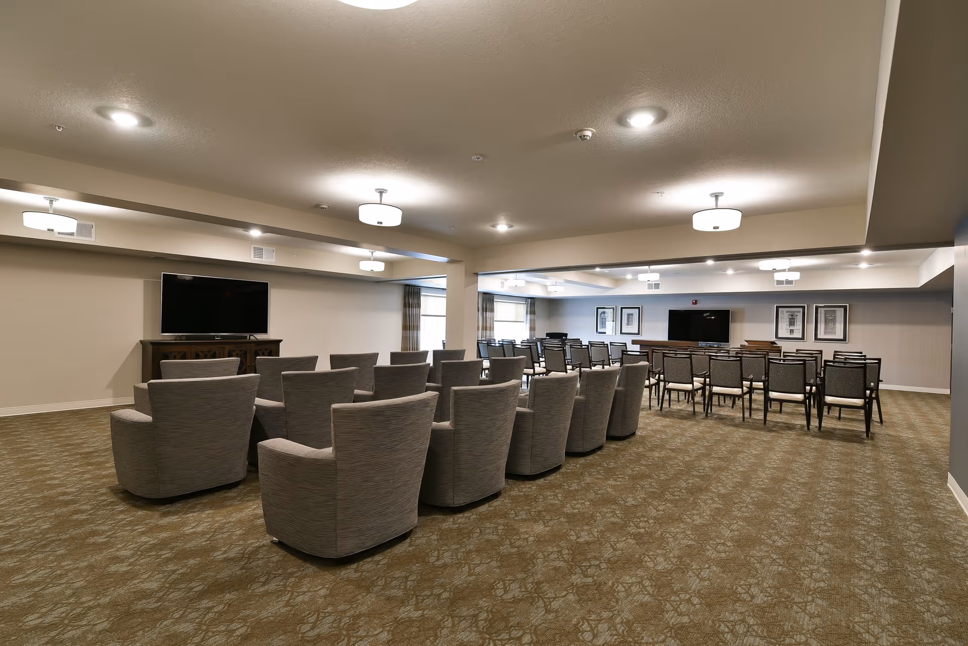 A large room with two sections of seating facing two flat-screen TVs mounted on the walls. The front section has cushioned armchairs arranged in rows, while the back section has dining-style chairs and tables. The room has beige walls, patterned carpet, ceiling lights, and framed artwork on the walls.