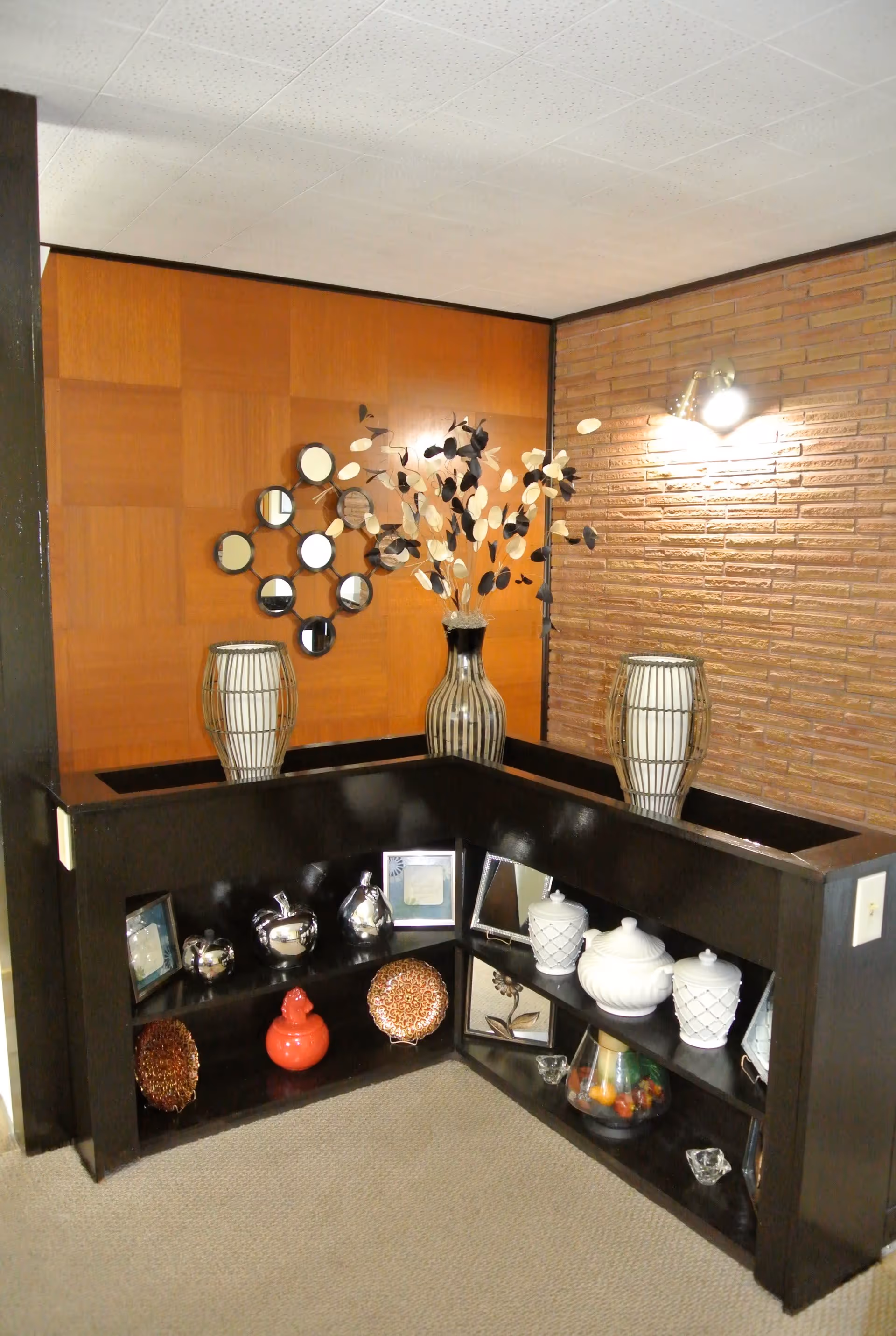 Corner shelf unit with decorative items including vases, framed pictures, ornamental apples, a red ceramic container, and a glass bowl with artificial fruit. The background features a wood-paneled wall and a brick wall with a wall-mounted light fixture.