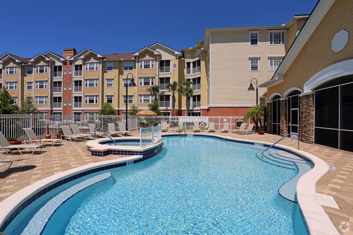 Outdoor swimming pool and lounge area in front of a multi-story yellow residential building under a clear blue sky.
