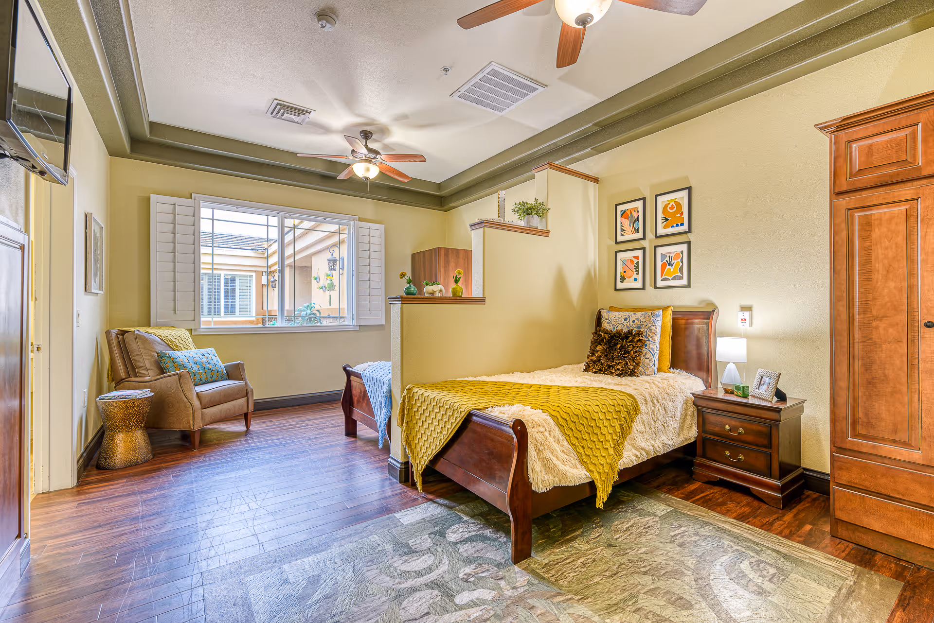 A bright and cozy bedroom in a senior living facility featuring a single wooden bed with yellow and beige bedding, a wooden nightstand with a lamp and picture frame, a comfortable armchair with cushions, a large window with white shutters, ceiling fans, and wooden flooring with a patterned rug.
