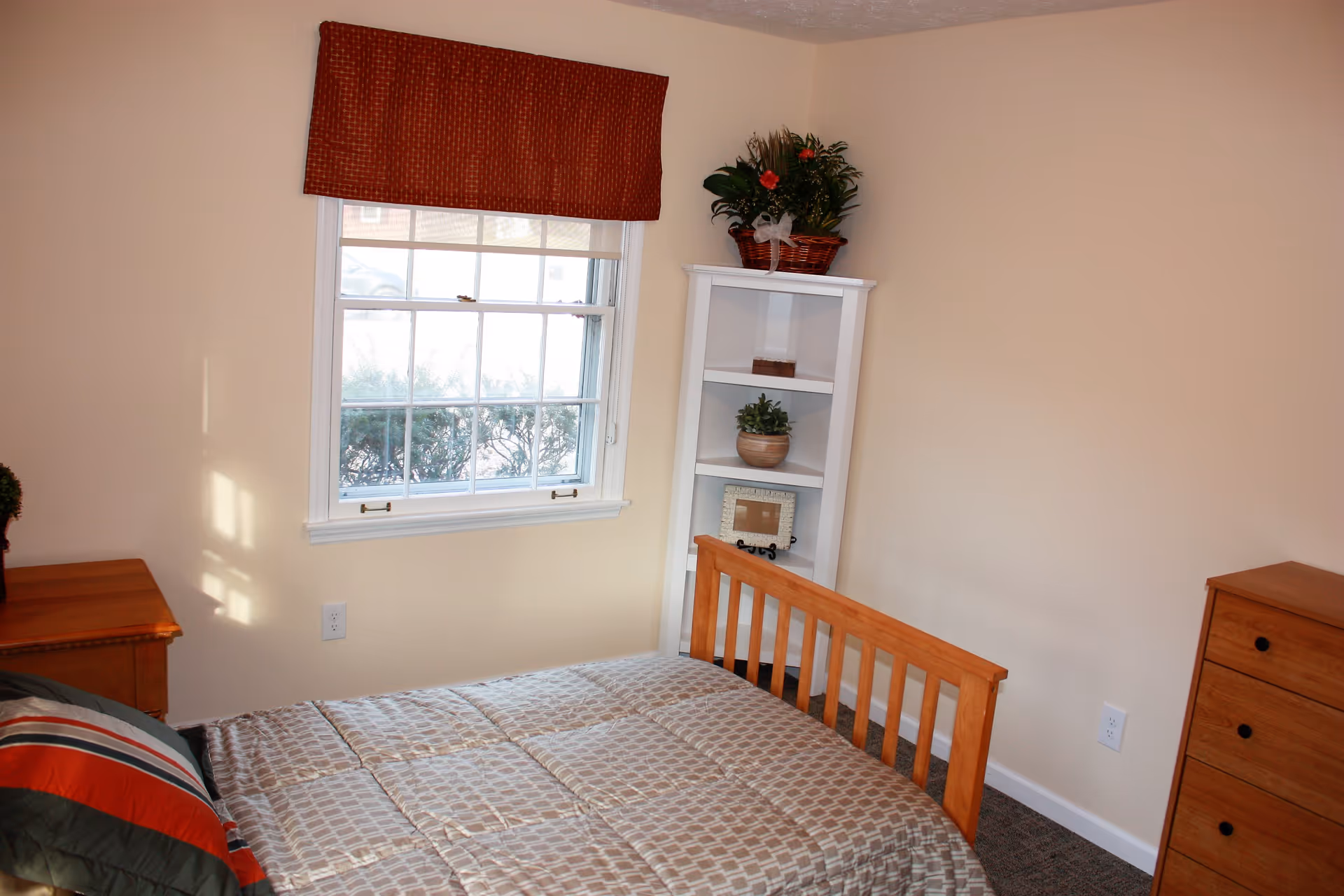 A small bedroom with a wooden bed frame and a patterned beige comforter. There is a window with a red valance letting in natural light. A white corner shelf holds a basket with flowers, a small potted plant, and a picture frame. A wooden dresser and nightstand are also visible.