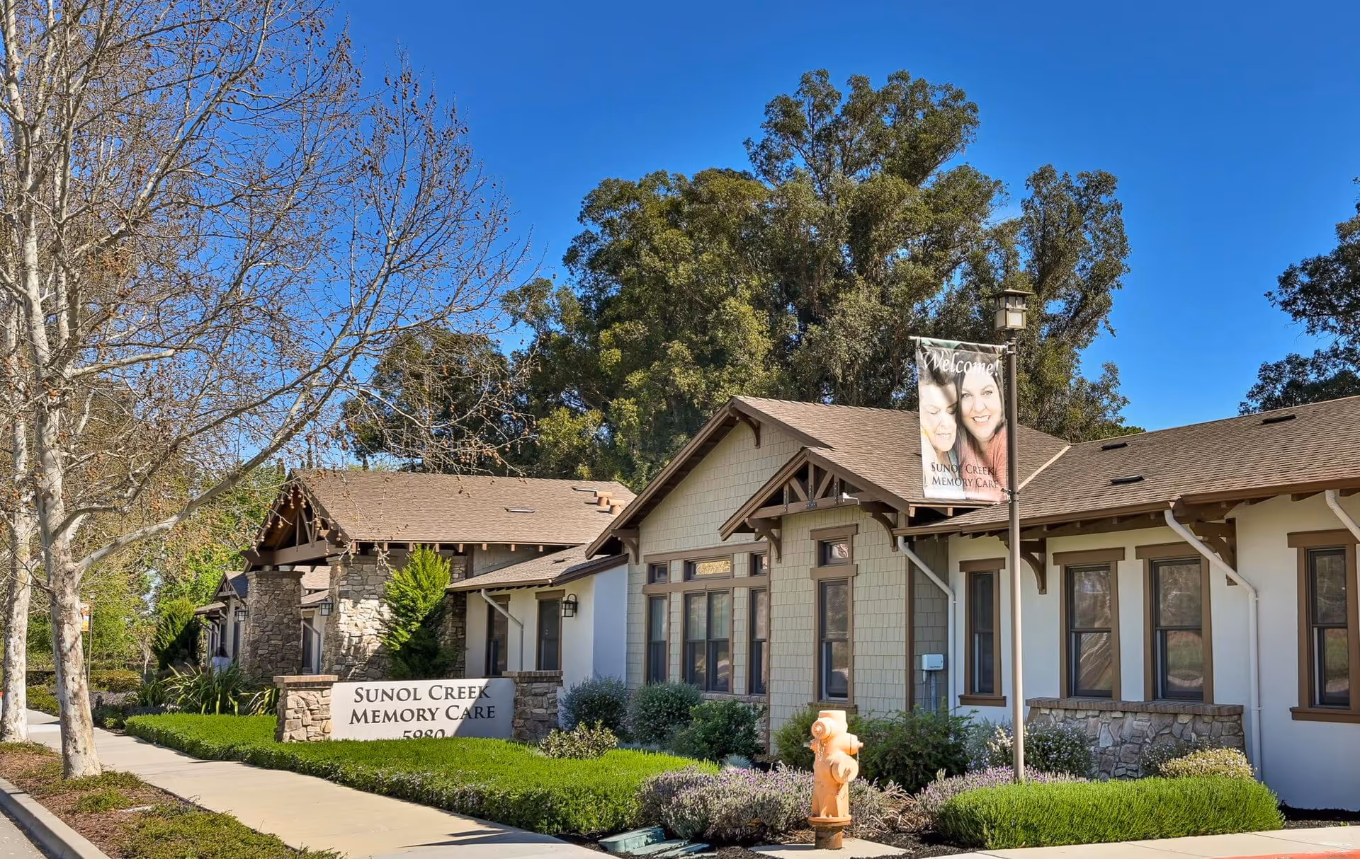 Exterior view of Sunol Creek Memory Care facility showing a single-story building with beige siding, brown trim, and a stone accent wall. There is a sidewalk and landscaped greenery including bushes and trees, with a clear blue sky overhead. A sign in front reads 'Sunol Creek Memory Care' and a banner on a lamp post says 'Welcome! Sunol Creek Memory Care' with images of two smiling women.