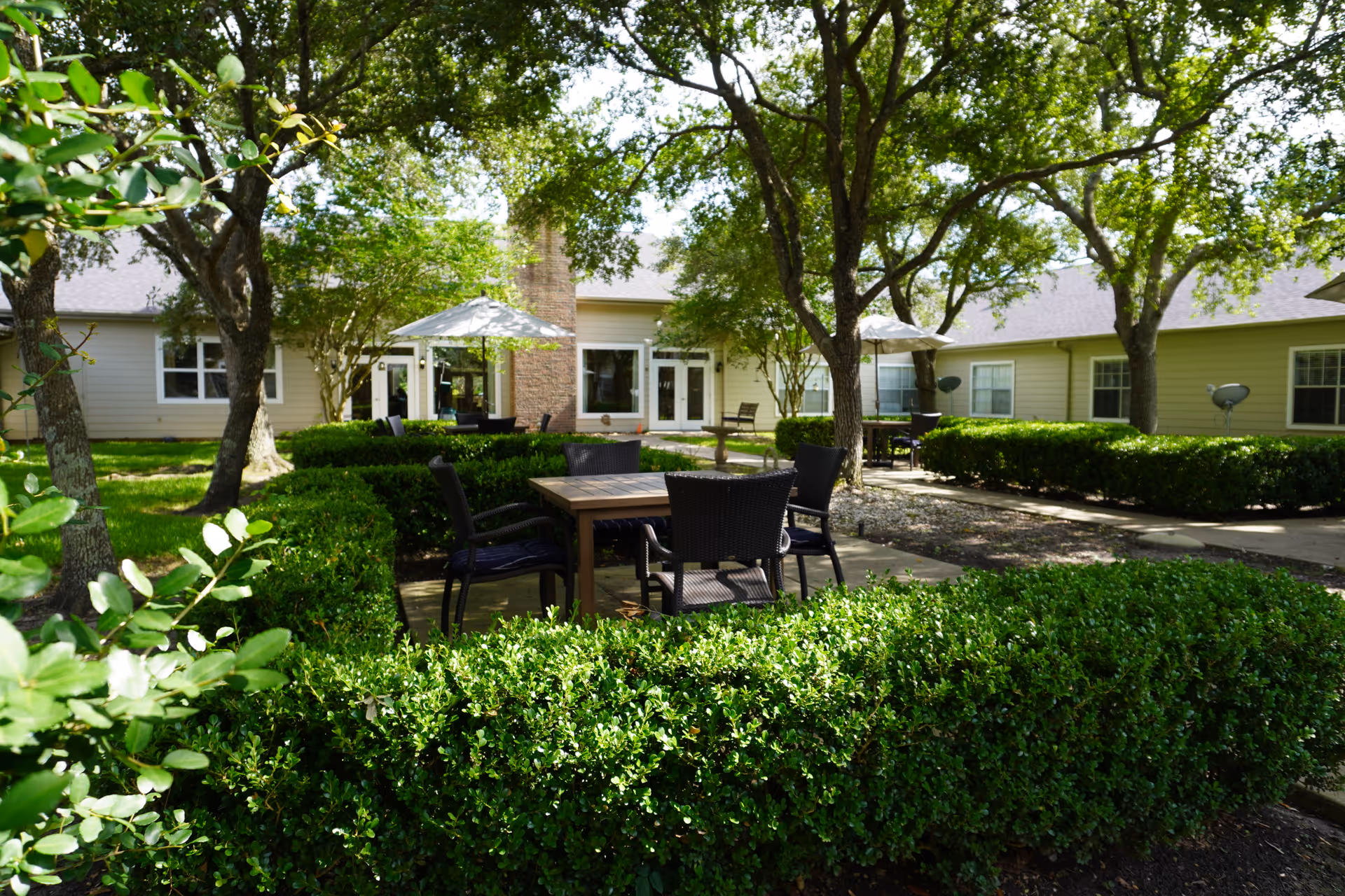 Outdoor courtyard area at Sodalis Texas City featuring green hedges, several trees providing shade, multiple patio tables with chairs, and umbrellas. The building with beige siding and windows surrounds the courtyard.