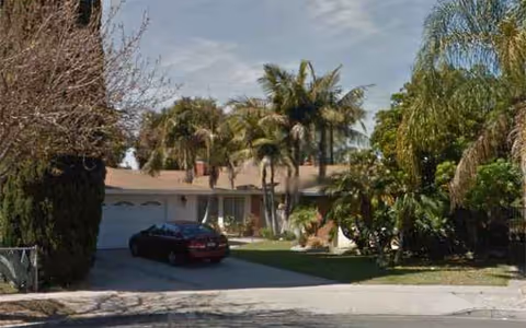 Front exterior of a single-story house with a driveway, parked car, and palm trees.