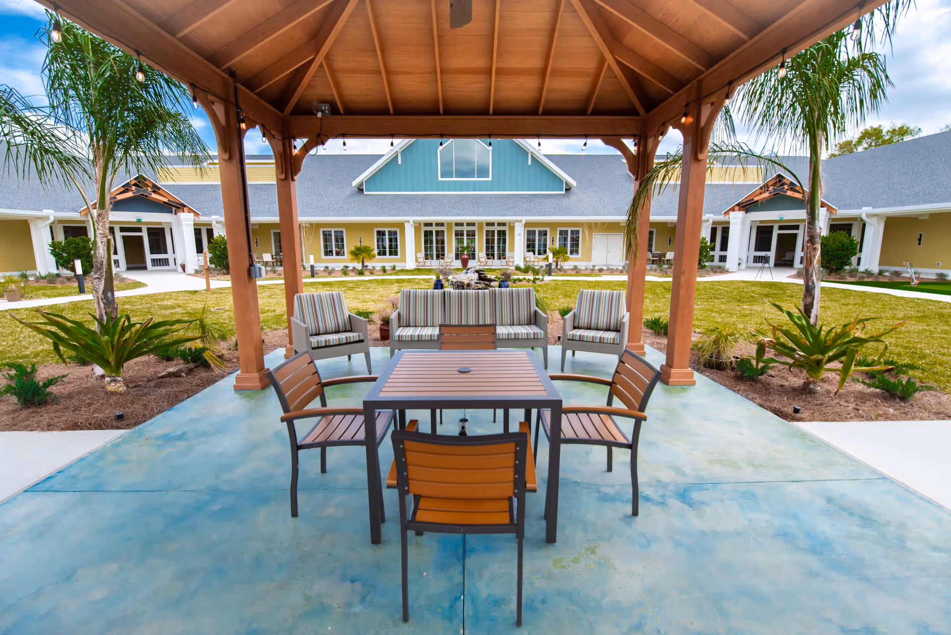 Covered outdoor patio with a table, chairs and cushioned seating facing a grassy courtyard and the facility building.