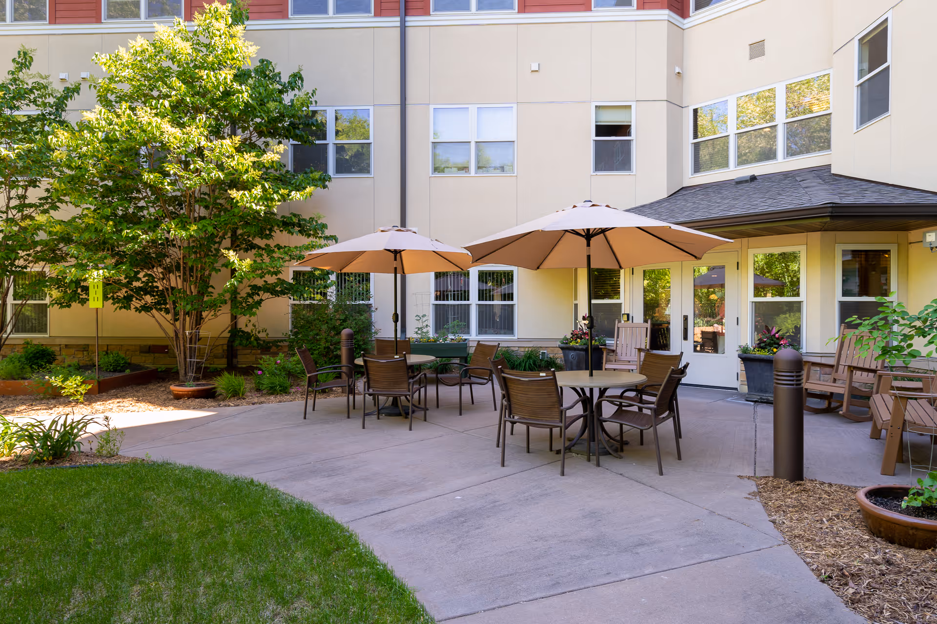 Outdoor patio area at The Glenn Minnetonka with round tables, chairs, and large beige umbrellas. The patio is surrounded by greenery, potted plants, and the exterior walls of the building with multiple windows and a door.