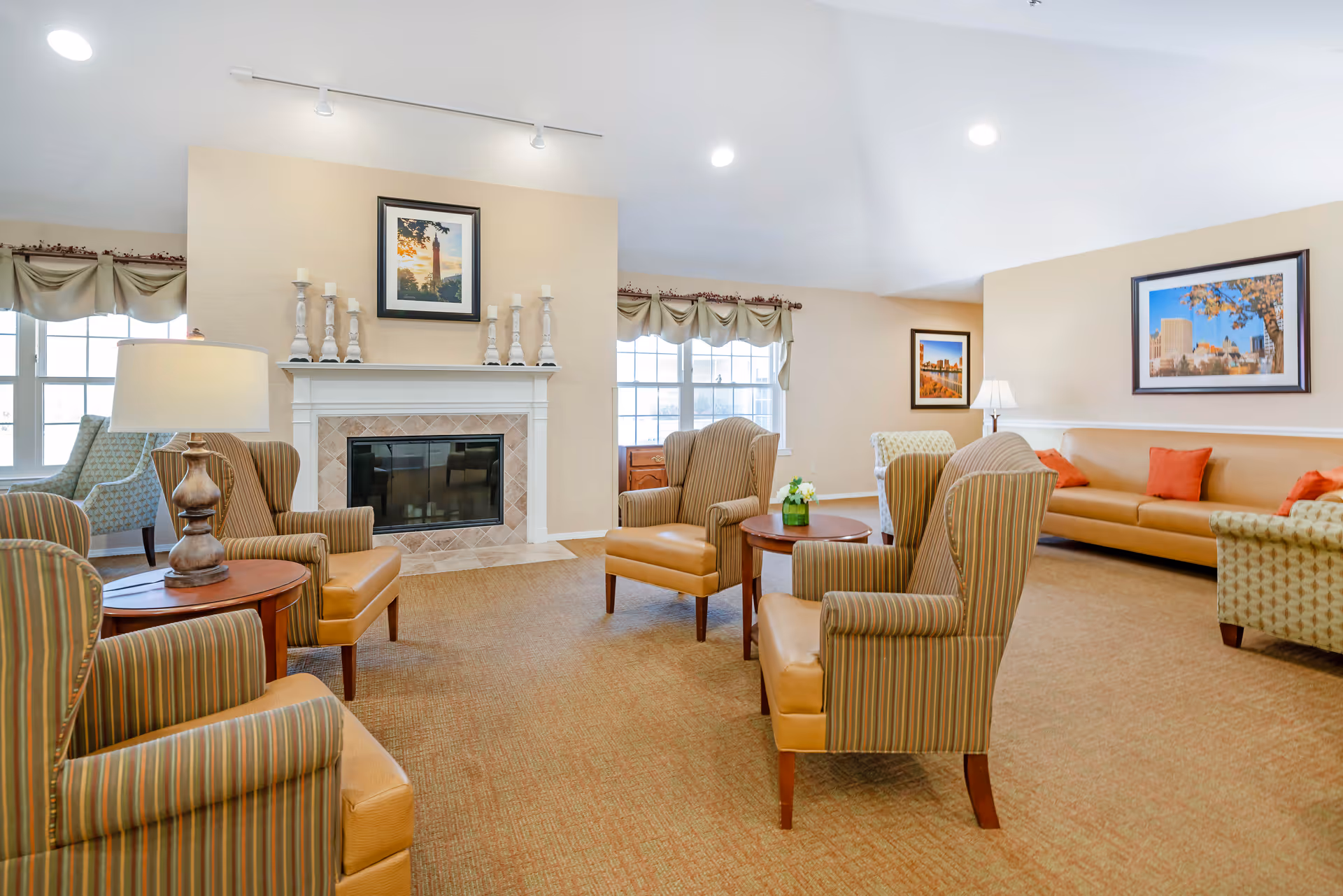 A bright and spacious living room area with several striped armchairs and a long beige sofa with orange cushions arranged around a fireplace. The fireplace mantel is decorated with white candlesticks and a framed picture above it. Large windows with beige curtains allow natural light to fill the room. There are framed pictures on the walls and a small round wooden table with a flower vase in the center.