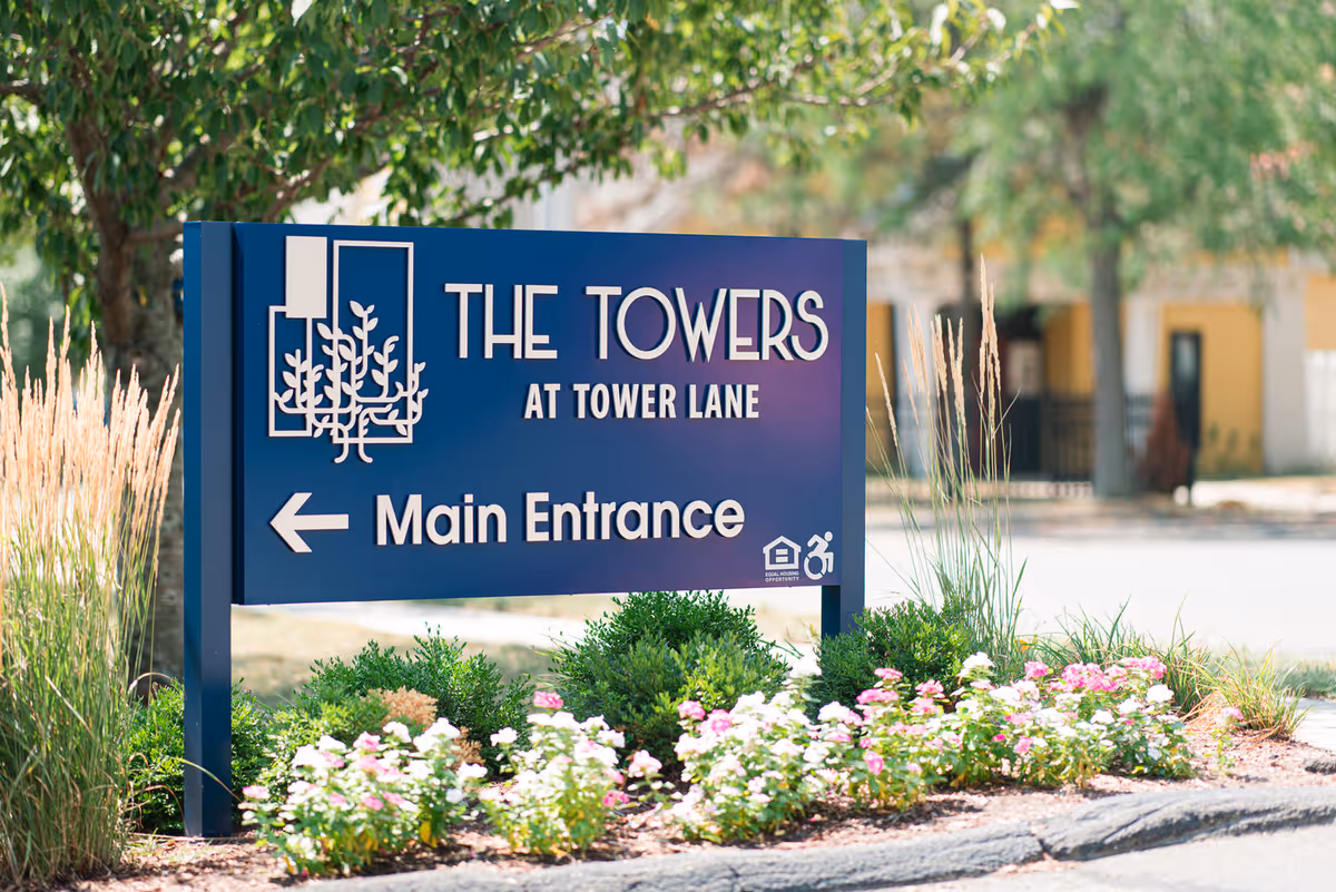 A blue sign for The Towers at Tower Lane indicating the main entrance with an arrow pointing left, surrounded by greenery and flowers with trees and a building in the background.