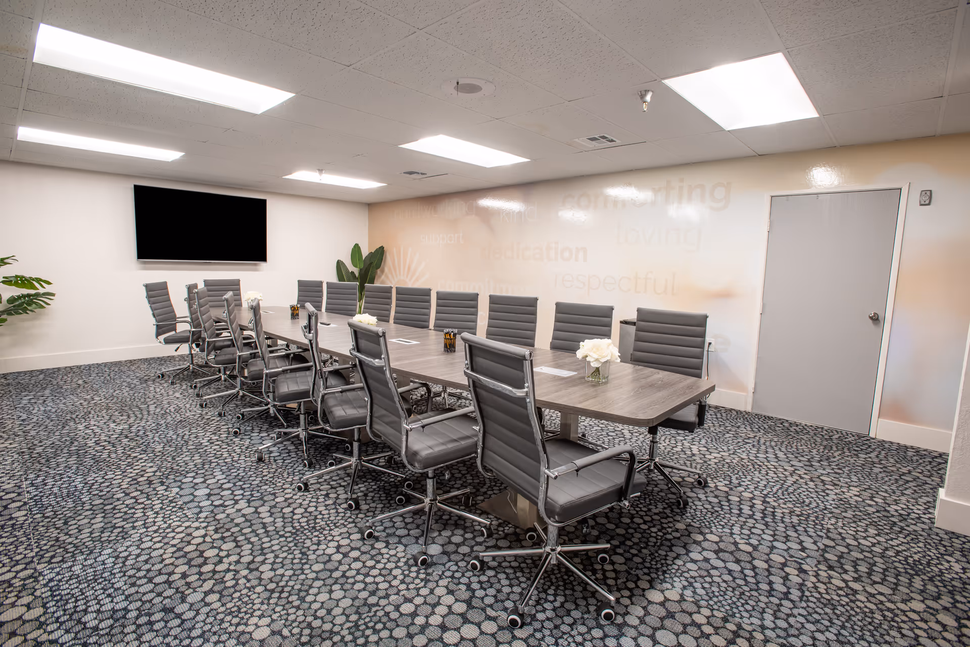 A modern conference room with a long rectangular table surrounded by gray office chairs on wheels. The room has a patterned carpet, a large flat-screen TV mounted on the wall, and some plants in the corners. The walls are light-colored with faint words like 'kind', 'comforting', 'loving', and 'respectful' visible on one side.