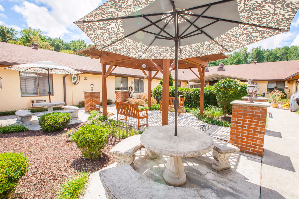 Outdoor courtyard area with stone tables and benches, large patterned umbrellas providing shade, wooden benches under a wooden pergola, surrounded by bushes and plants, with a single-story building in the background under a partly cloudy sky.
