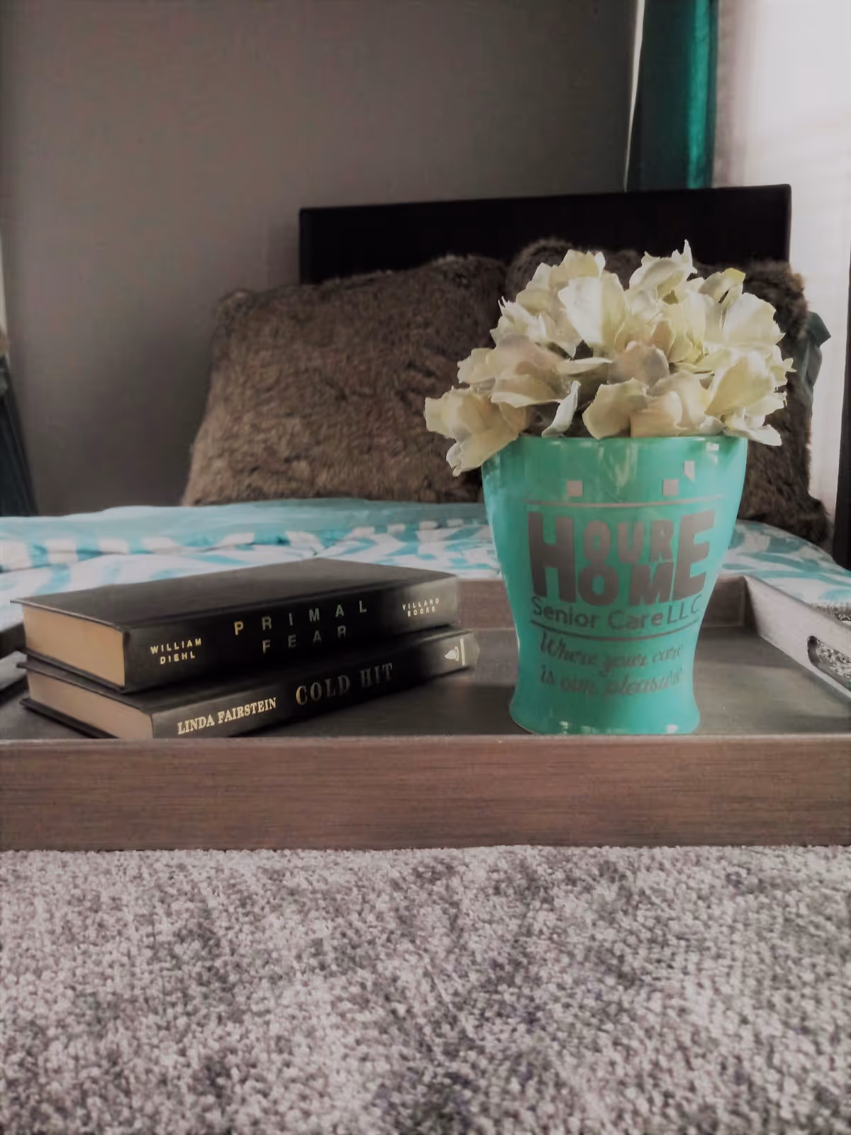 A wooden tray on a bed with a teal flower pot containing white flowers and two stacked books titled 'Primal Fear' and 'Cold Hit'. The flower pot has the text 'Your Home Senior Care LLC' and 'Where your care is our pleasure' printed on it. The bed has a fluffy brown pillow and a teal and white patterned blanket.