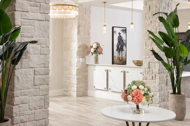 Bright interior space with light-colored stone pillars, a round white table with a floral arrangement, large green potted plants, a white cabinet with decorative items including a vase of flowers, and a framed painting of a person on horseback on the wall.