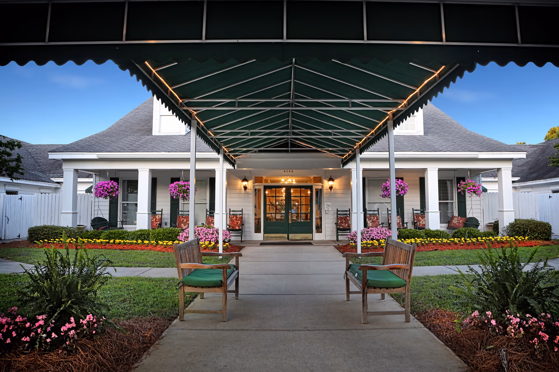 Entrance to a senior care facility with a covered walkway, two wooden benches with green cushions, hanging flower baskets, and a well-maintained garden with colorful flowers and shrubs.