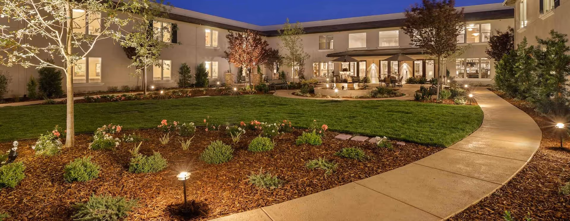 Evening view of a well-lit courtyard garden at a senior living facility with a curved concrete pathway, green lawn, flower beds, trees, and a seating area with tables and umbrellas near the building.