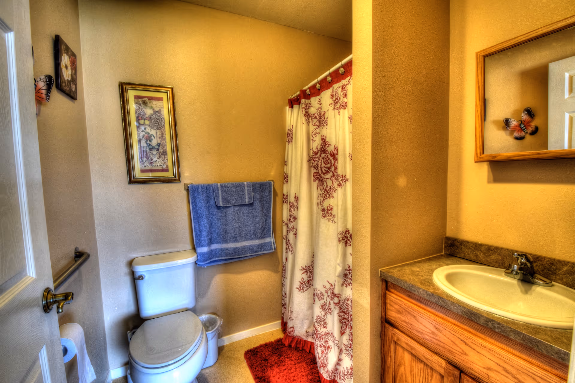 A small bathroom with beige walls featuring a white toilet with a blue towel hanging above it, a shower with a white curtain decorated with red floral patterns, a red bath mat on the floor, a wooden vanity with a sink and faucet, a mirror above the sink, and wall decorations including framed artwork and butterfly ornaments.