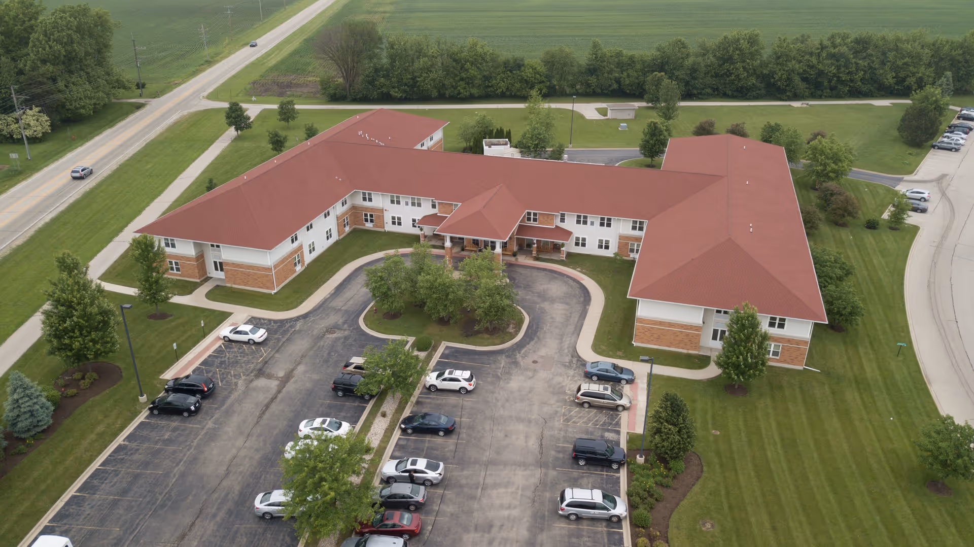 Aerial view of a large senior living facility building with a red roof and white walls, surrounded by green lawns and trees. The building is shaped like a U with a covered entrance in the center. There is a parking lot with several cars parked in front of the building, and a road runs alongside the property.