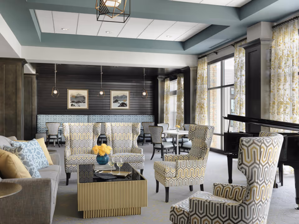 A bright and elegant living room area in Rolling Green Village featuring patterned armchairs, a gray sofa with blue and yellow pillows, a black grand piano, and large windows with floral curtains allowing natural light to fill the space. The room also includes a black coffee table with a vase of yellow flowers and wine glasses, pendant lights, and framed artwork on a dark wood-paneled wall.