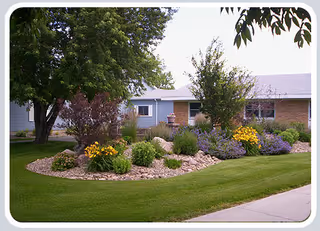 Manicured lawn with a rock-edged flowerbed of shrubs, yellow blooms and trees in front of a single-story brick and siding building.