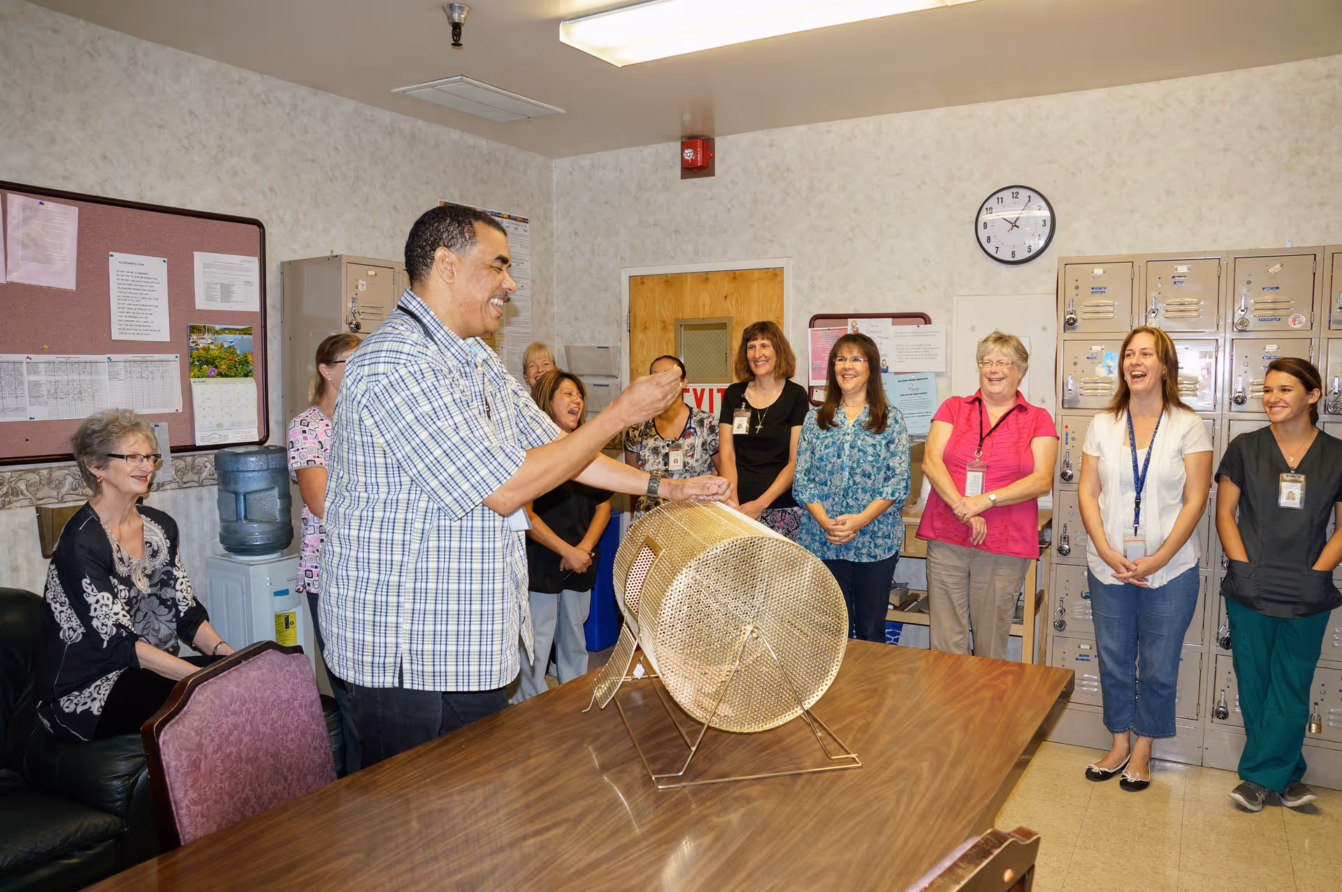 A group of people standing and sitting in a room with lockers and a bulletin board. A man in a plaid shirt is turning a large metal raffle drum on a table while others watch and smile.