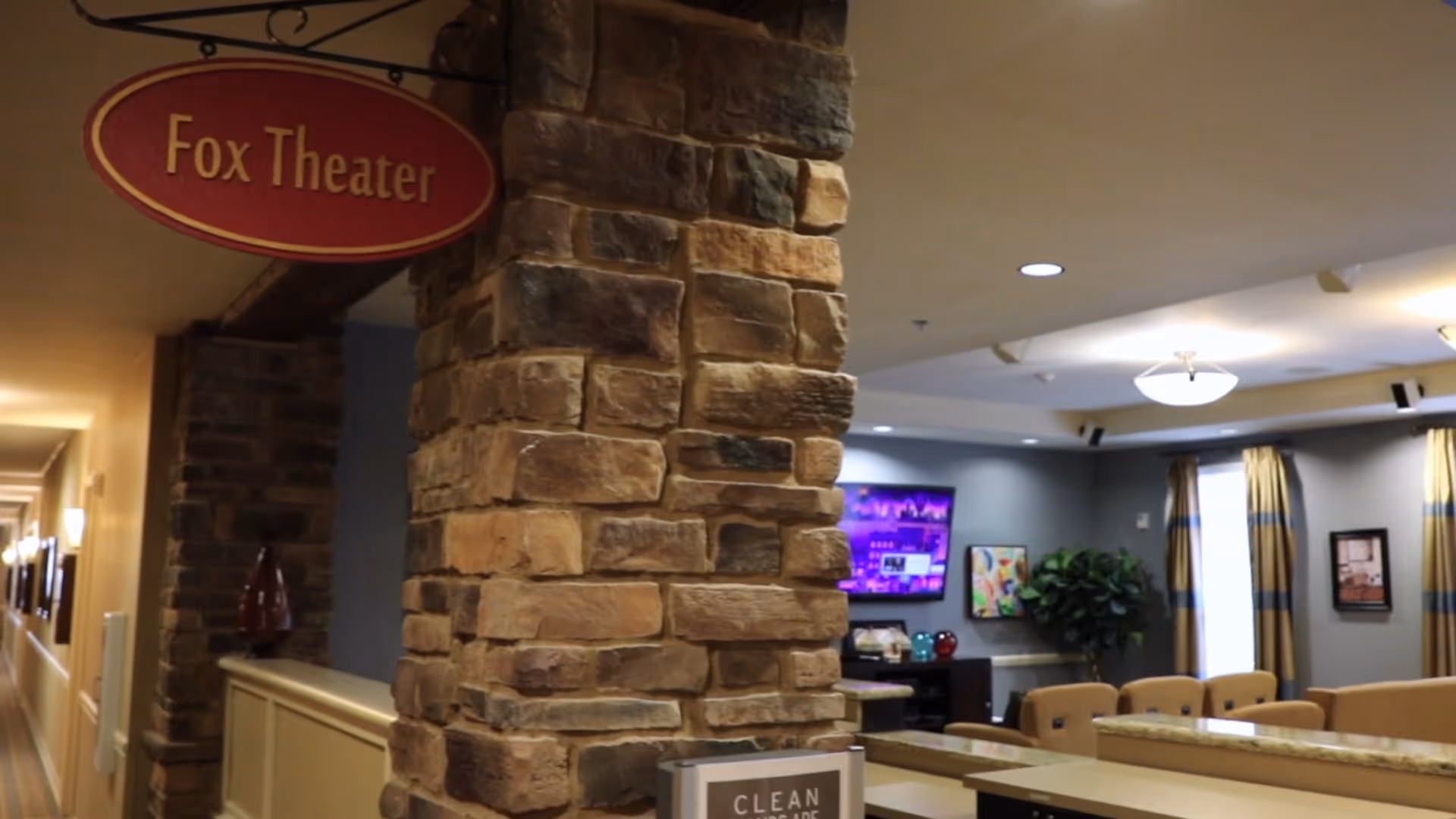 Interior view of a senior living facility showing a stone pillar with a hanging red sign that reads 'Fox Theater'. In the background, there is a lounge area with beige chairs, a television mounted on a blue-gray wall, framed artwork, curtains on windows, and ceiling lights.