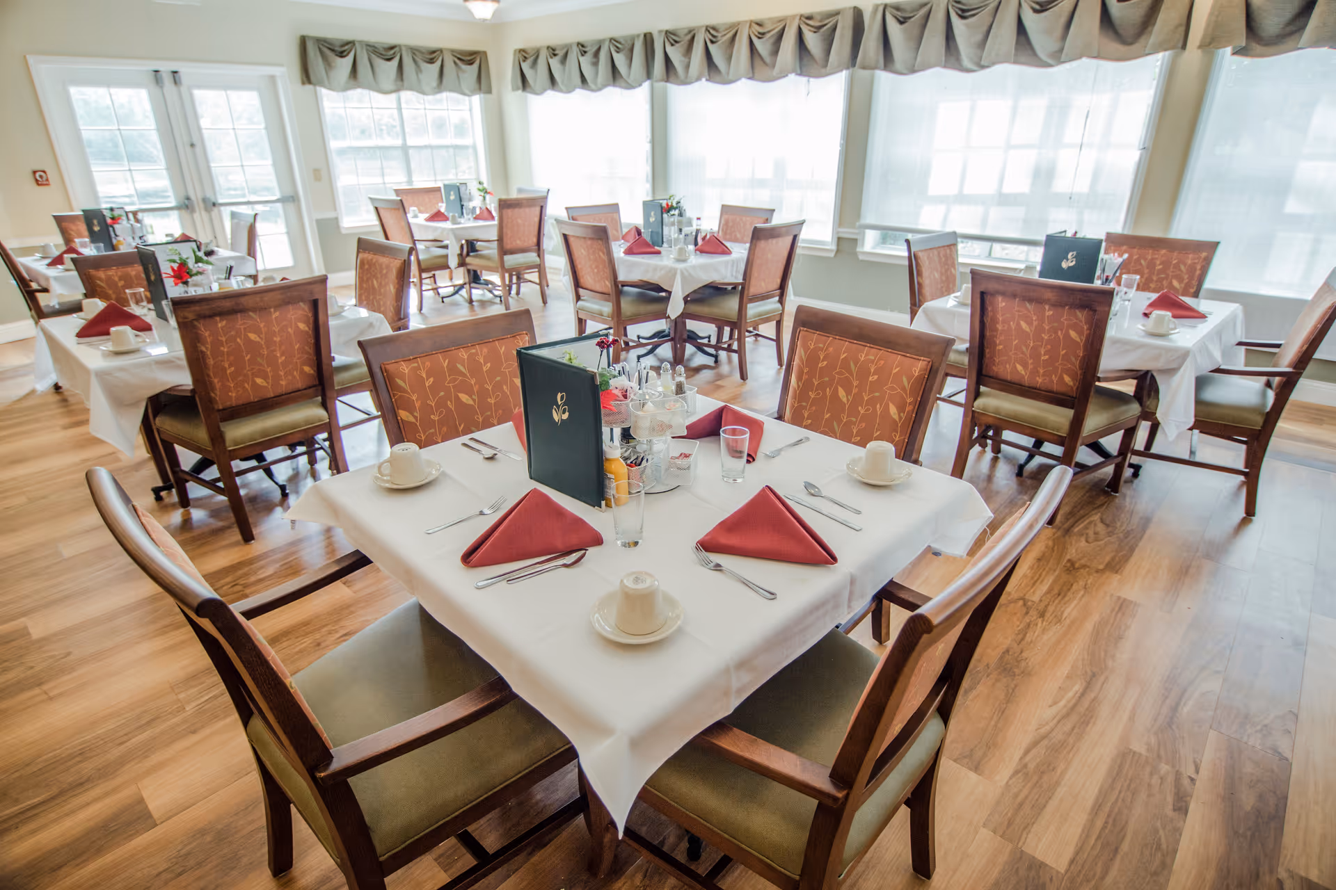 Bright dining room with multiple set tables, wooden chairs, and folded red napkins by large windows.