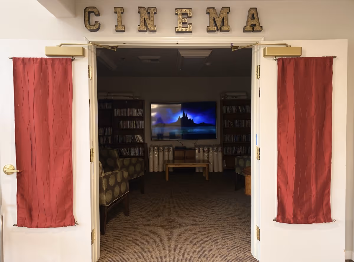 Open double doors with red curtains on each door panel leading into a small cinema room. Inside the room, there are patterned armchairs, bookshelves filled with DVDs or books, a wooden coffee table, and a large flat-screen TV mounted on the wall displaying a castle scene. Above the doors, large decorative letters spell out 'CINEMA'.