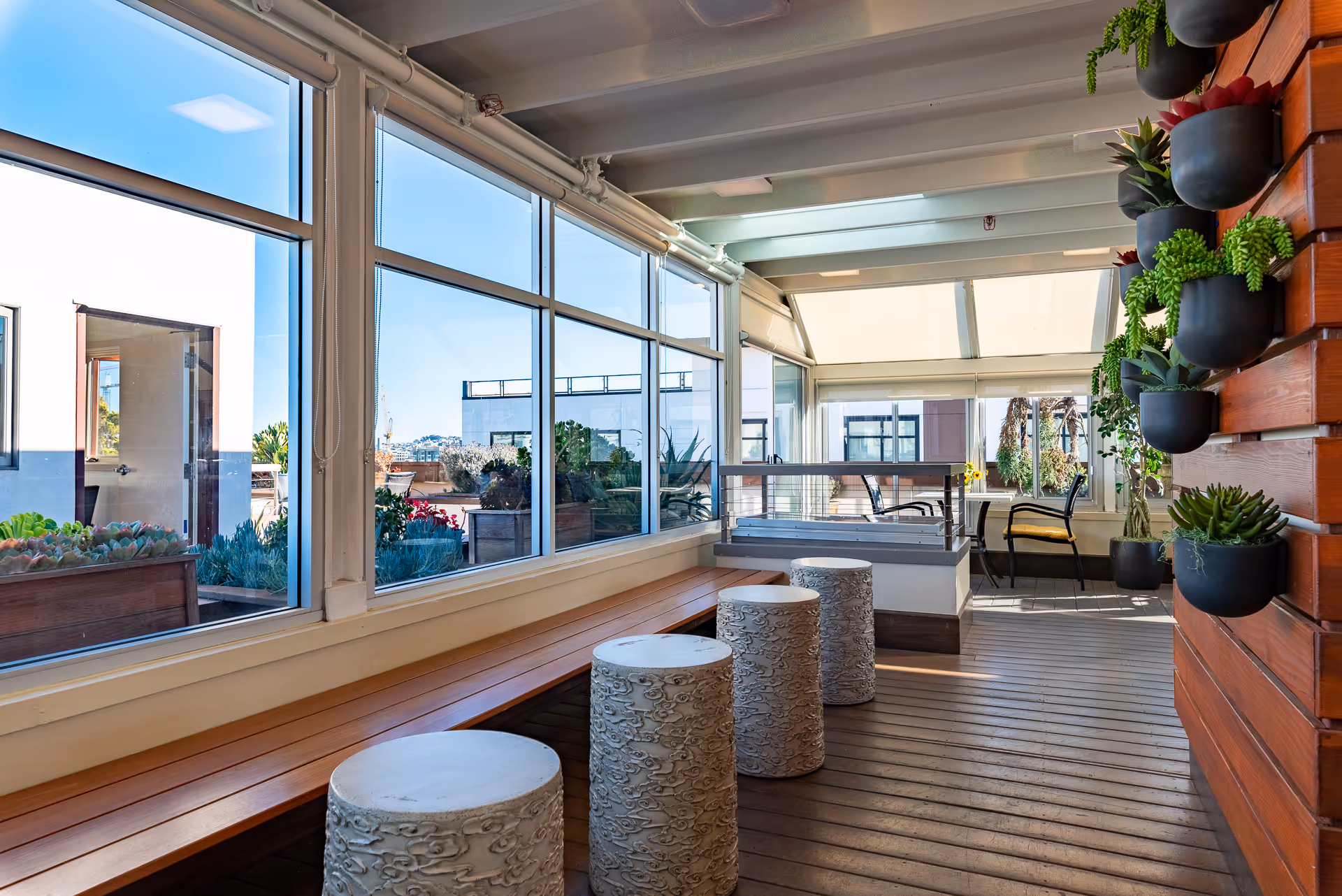 Bright enclosed sunroom-style common area with large windows, a long wooden bench, decorative stools and wall-mounted potted plants.