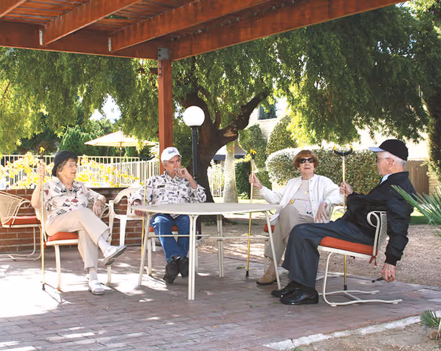 Four elderly people sitting around a round outdoor table under a wooden pergola in a garden area. They are seated on metal chairs with cushions, holding walking sticks, and appear to be engaged in conversation. There are trees, bushes, and a fence in the background.
