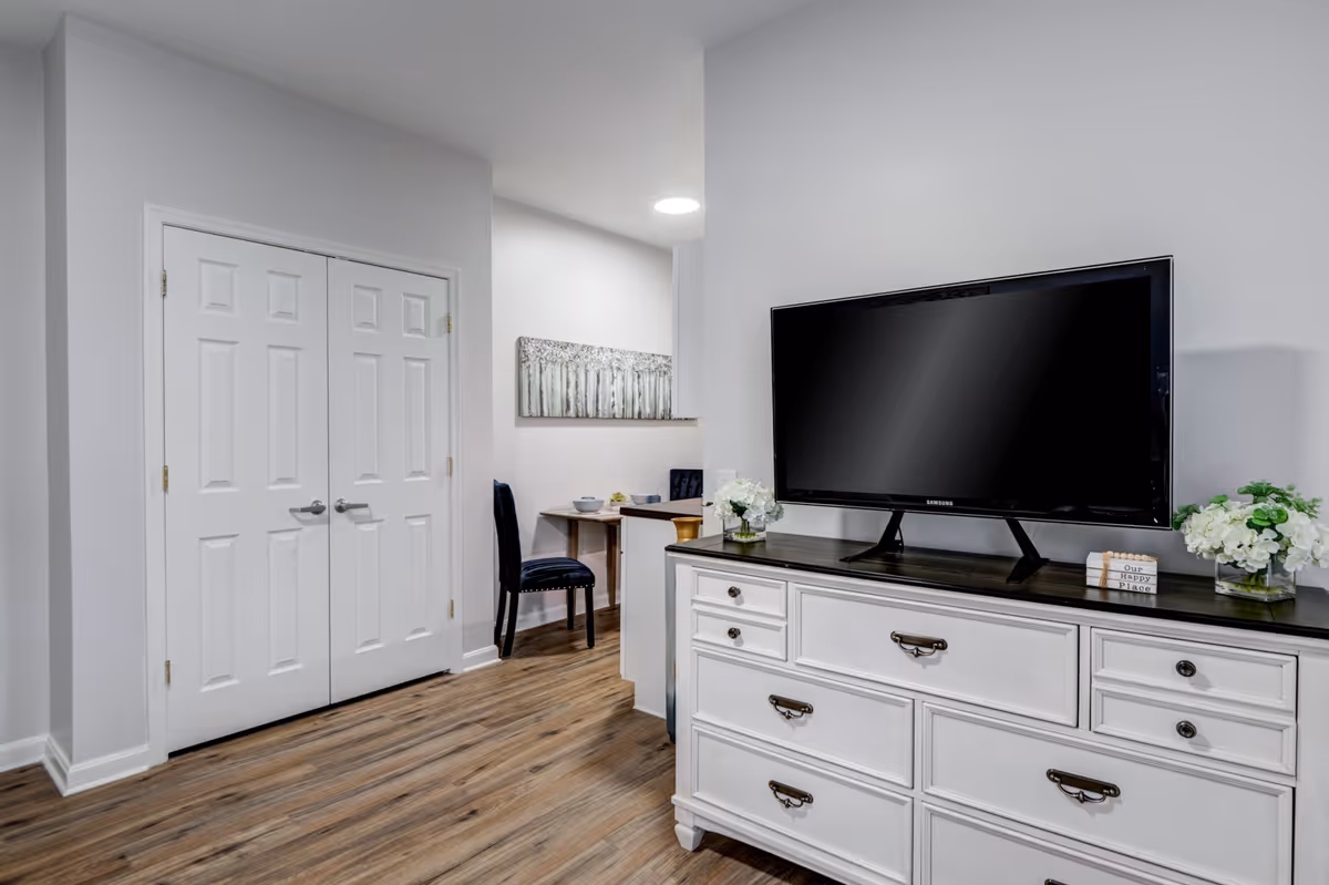 A bright interior room featuring a white dresser with a black top, a large flat-screen TV on the dresser, and two vases with white flowers on either side. To the left, there is a double-door closet and a small dining area with a table and two chairs. The room has wood flooring and light gray walls.