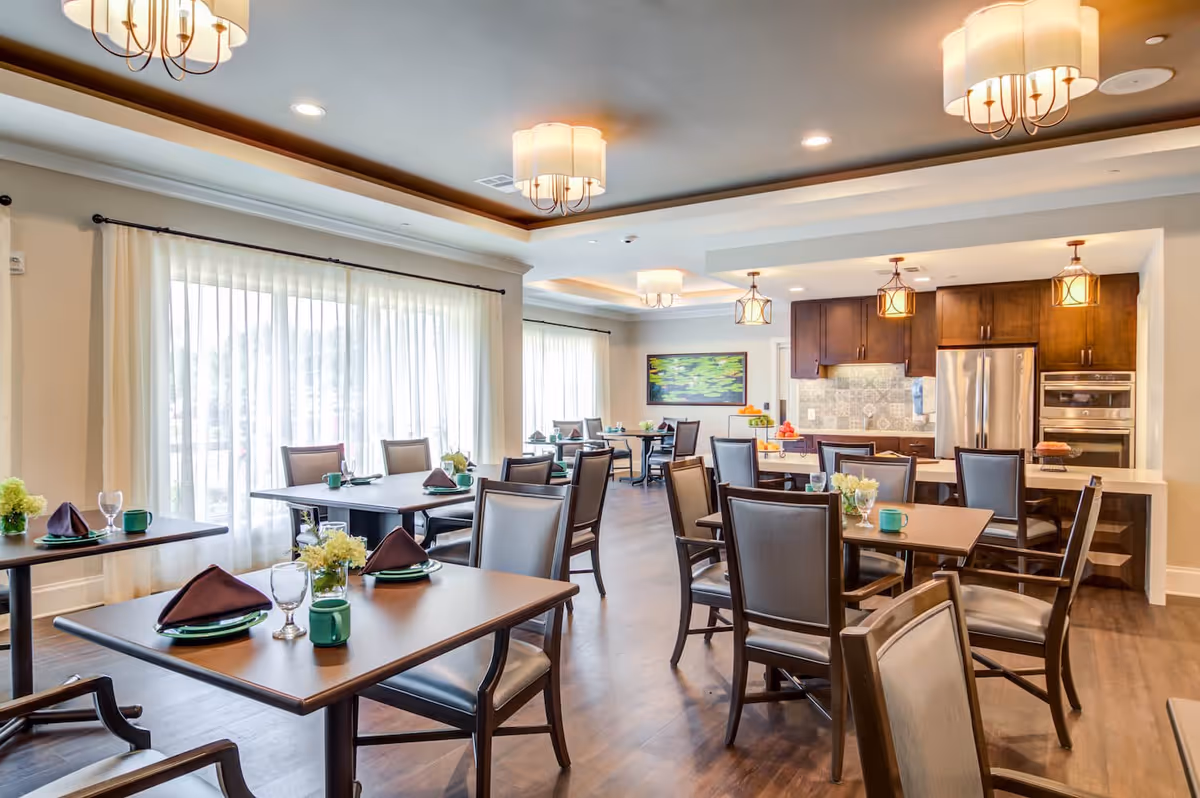 A bright and spacious dining room with multiple wooden tables and chairs arranged neatly. Each table is set with green plates, brown napkins, green mugs, and clear glasses. The room features large windows with sheer white curtains allowing natural light to fill the space. The ceiling has recessed lighting and decorative hanging light fixtures. In the background, there is a modern kitchen area with stainless steel appliances and wooden cabinets.