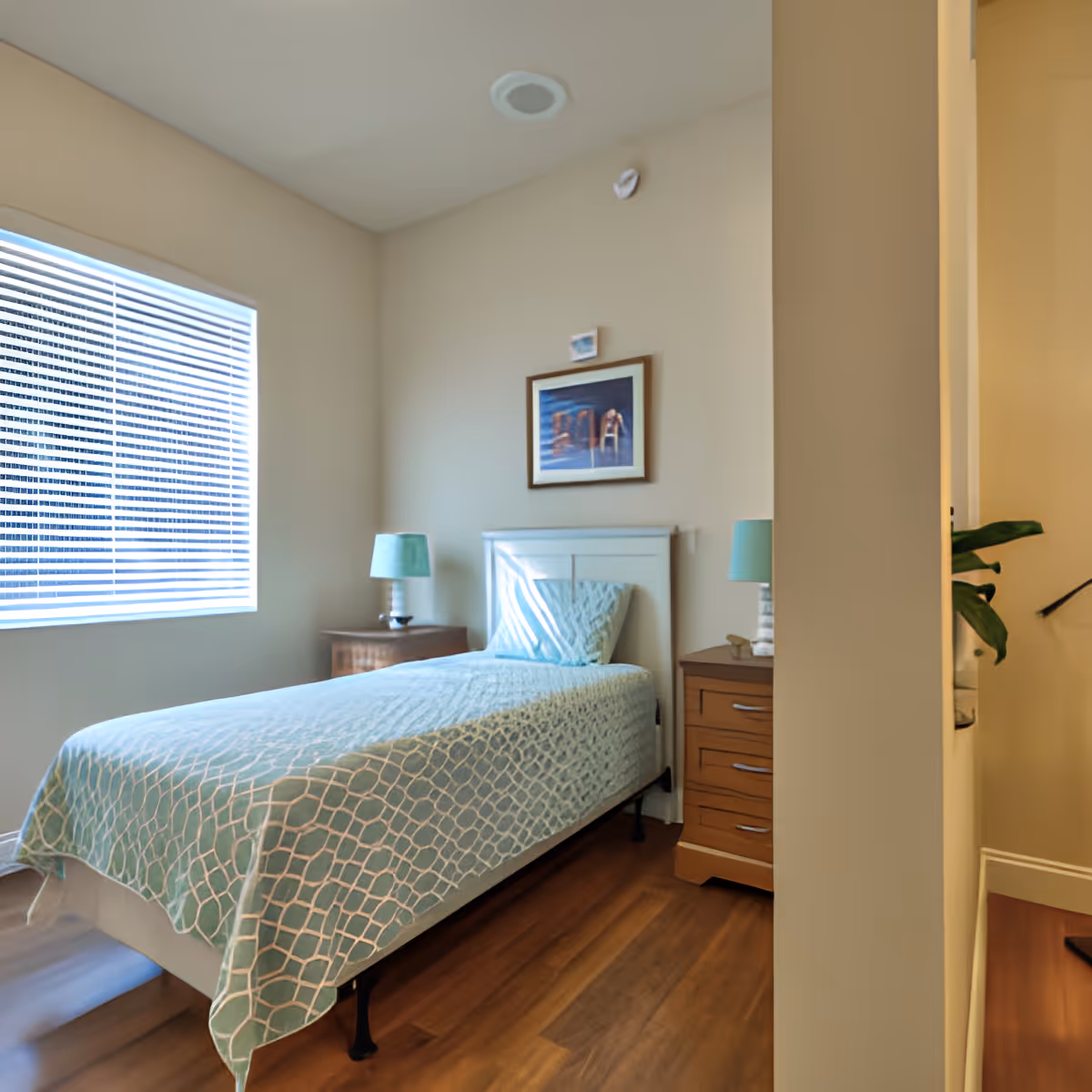 Sunny small bedroom with a single bed, two bedside tables and lamps, a window with blinds, framed artwork above the headboard, and wood flooring.