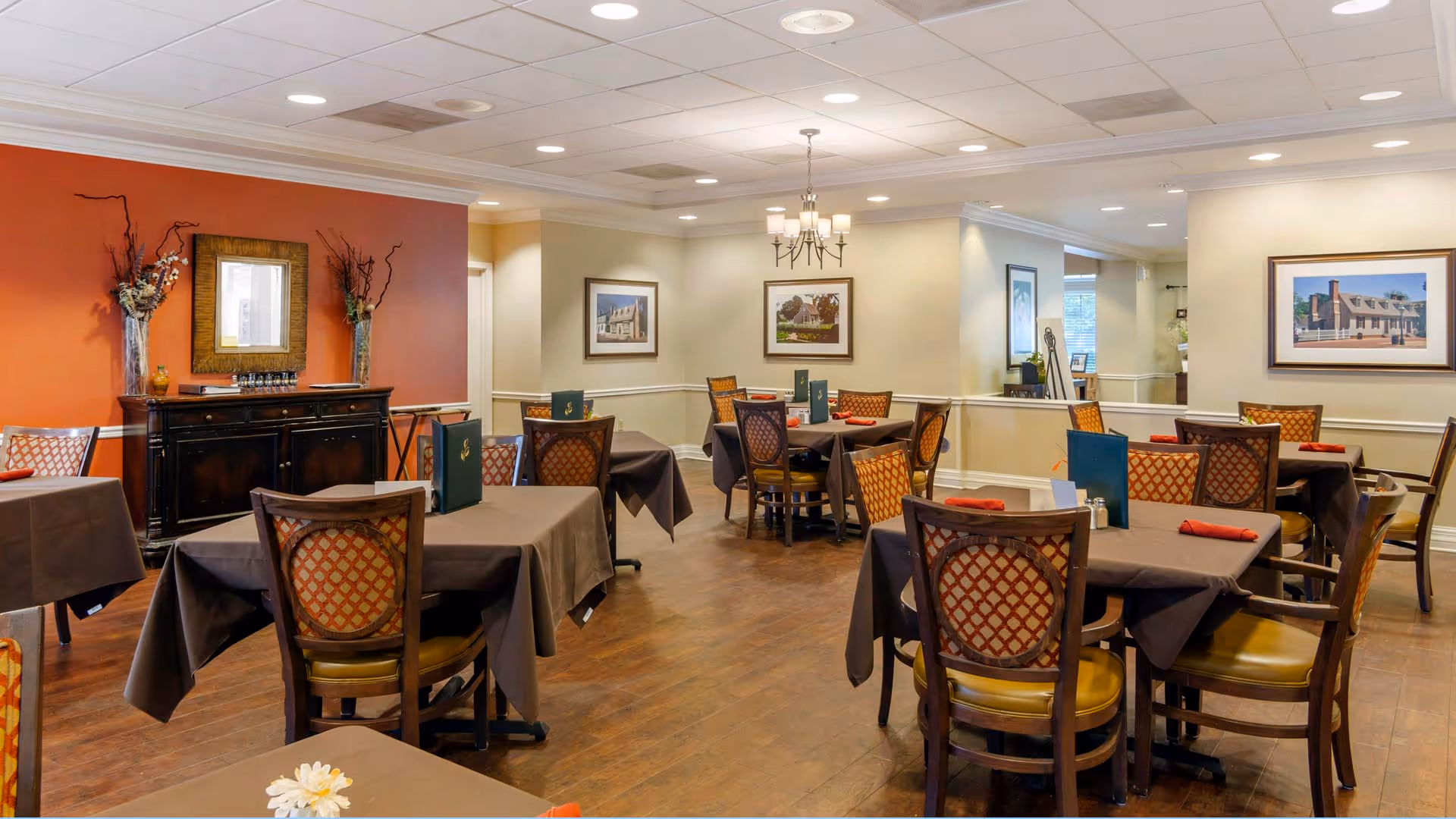 A dining room in a senior living facility with several tables covered in brown tablecloths, each set with menus and red napkins. The room has wooden chairs with patterned upholstery, warm lighting, framed pictures on the walls, and decorative vases with dried branches on a sideboard against a red accent wall.