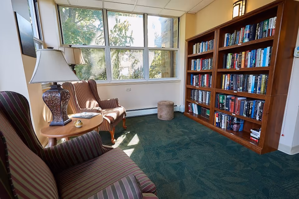 Sunlit reading nook with upholstered armchairs, a side table and lamp beside large windows and a filled wooden bookshelf.
