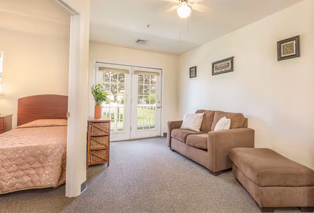 A cozy living area with a brown sofa and matching ottoman, beige walls decorated with framed artwork, and a ceiling fan with a light. Adjacent to the living area is a bedroom with a bed covered in a beige quilt. French doors with glass panes open to a green outdoor space.