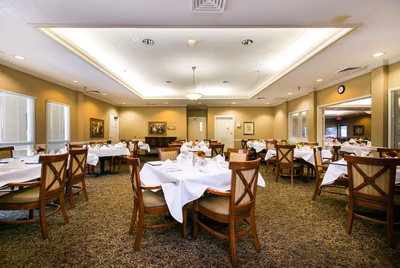 Well-lit formal dining room with multiple round tables set with white tablecloths and wooden chairs.