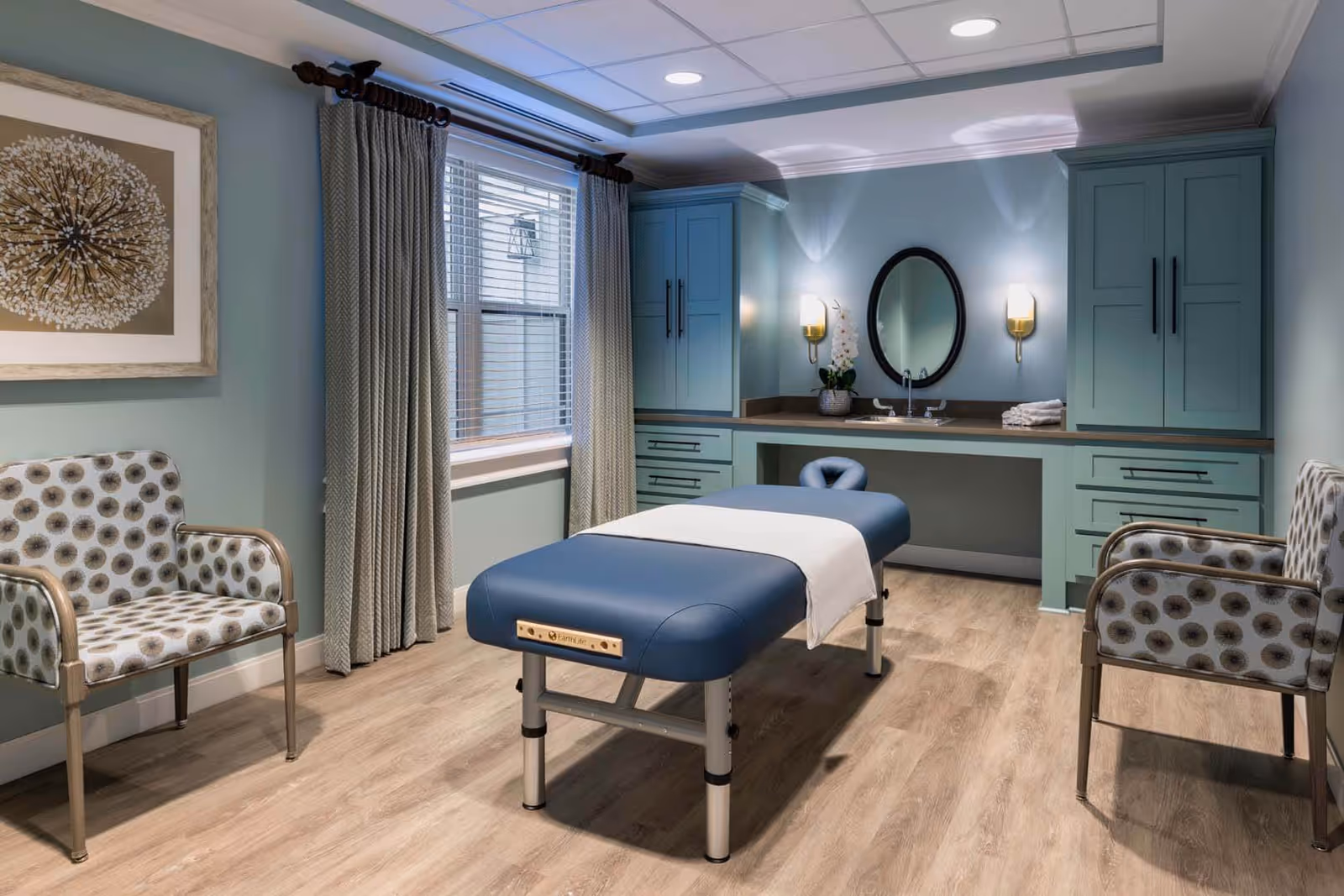 A well-lit room with a blue massage table in the center, flanked by two patterned armchairs. The room has light wood flooring, blue cabinetry with a countertop and sink, an oval mirror, wall sconces, a window with curtains, and a framed artwork on the wall.