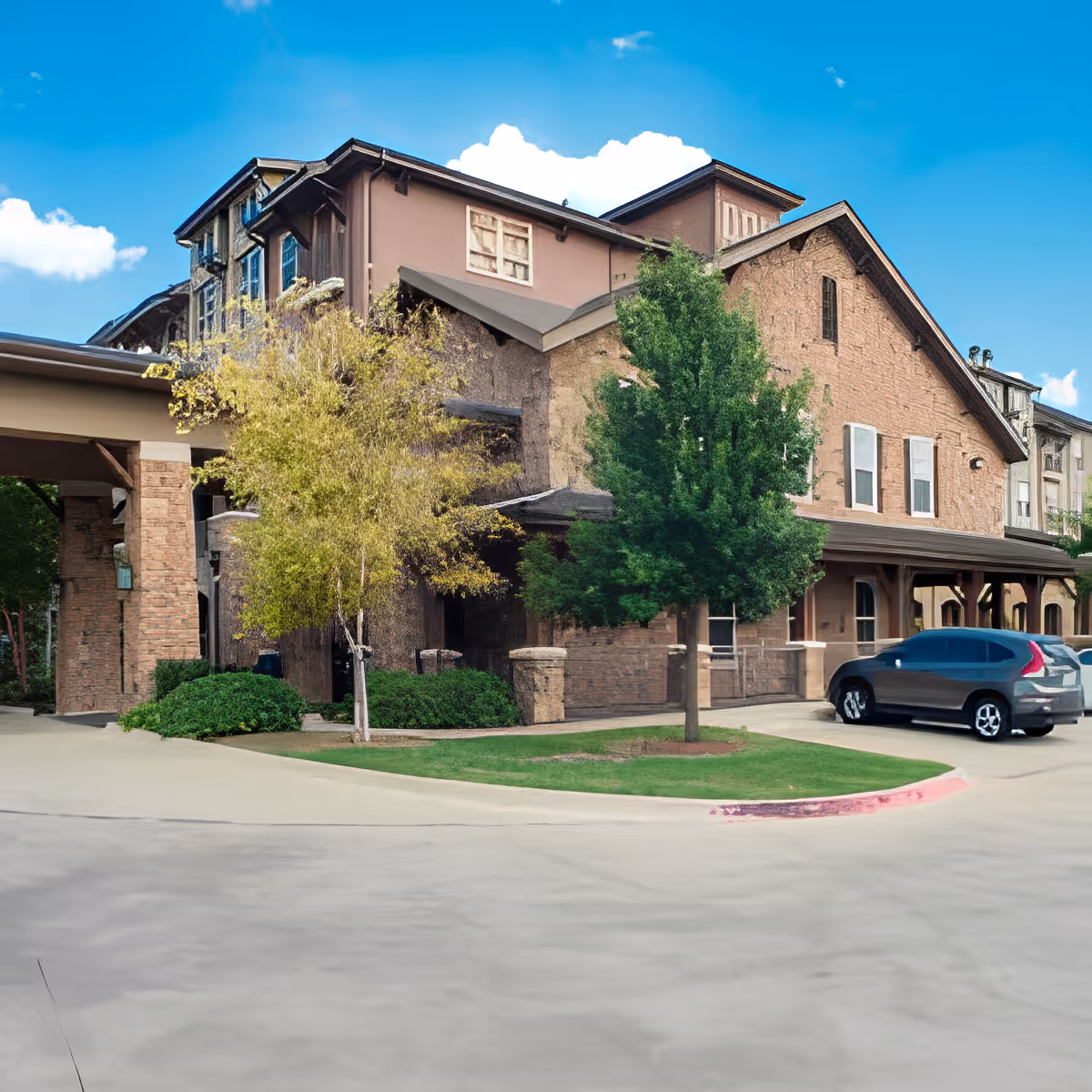 Exterior view of a multi-story senior living facility building with a stone and brick facade, surrounded by trees and greenery under a blue sky with clouds. There are parked cars near the entrance.