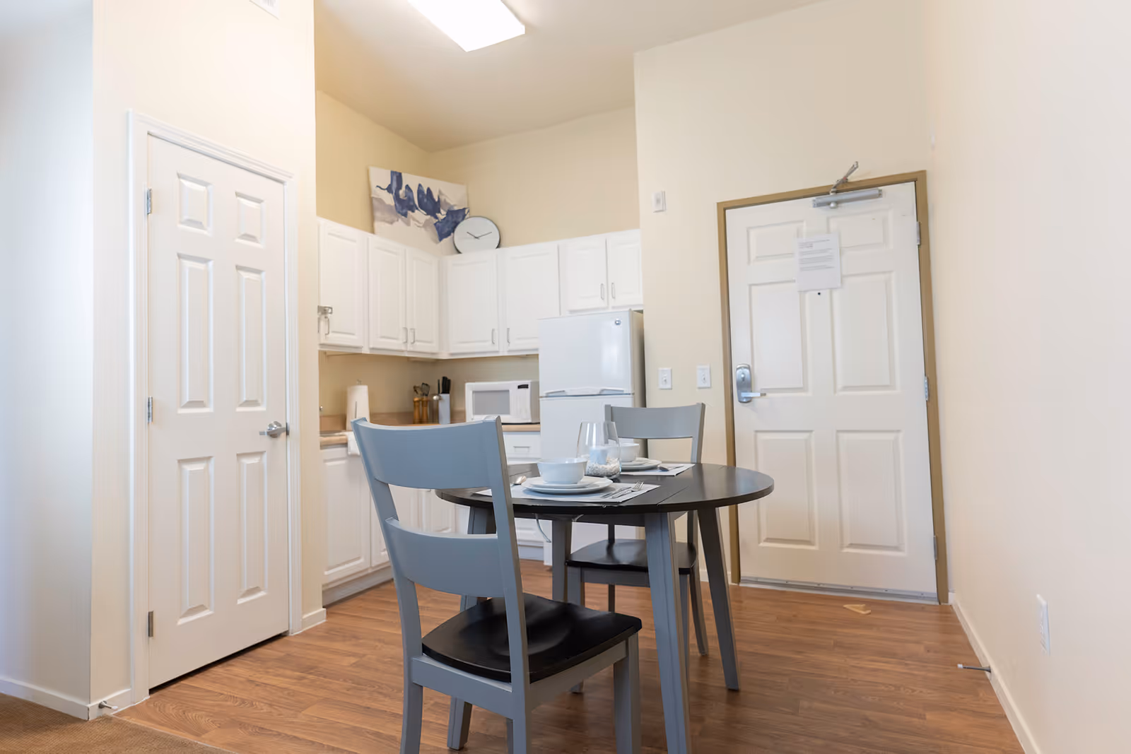 A small dining area in an assisted living facility with a round black table set for two with plates, bowls, and glasses. Two gray chairs are positioned around the table. In the background, there is a compact kitchen with white cabinets, a refrigerator, a microwave, and a countertop with utensils. A white door is visible on the right side, and another door is on the left. The room has light-colored walls and wood flooring.