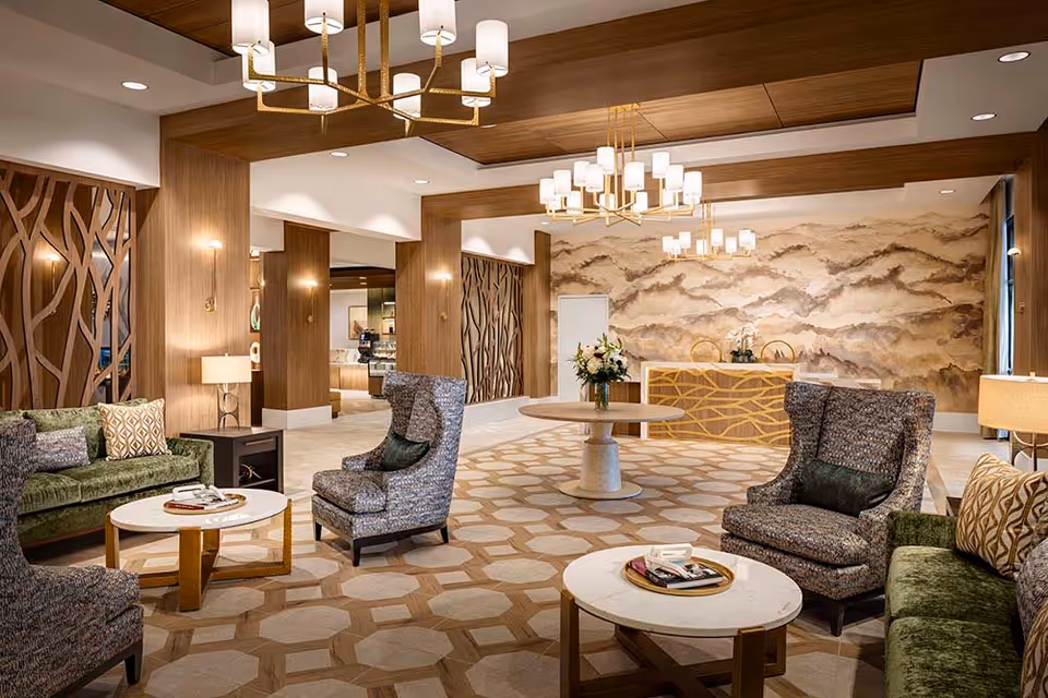 A spacious and elegantly decorated living room area with patterned carpet, two green sofas, two gray armchairs, and round white marble coffee tables. The room features wooden paneling, modern chandeliers, and a decorative wall with a nature-inspired design behind a reception desk.