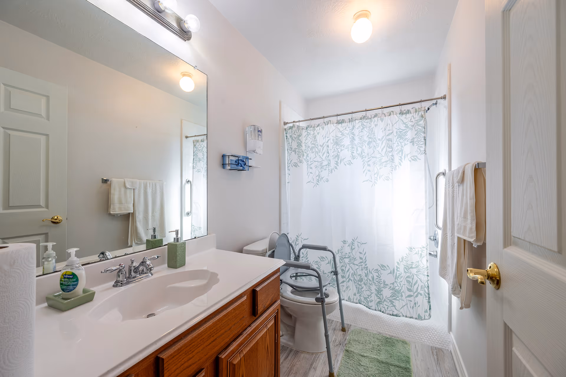 A clean and bright bathroom with a white sink countertop, wooden cabinet, large mirror, and a toilet equipped with a safety frame. There is a shower with a white curtain featuring a green leafy pattern, a green bath mat on the floor, and white towels hanging on the wall. The bathroom has light-colored walls and flooring, with a soap dispenser and hand sanitizer on the sink.