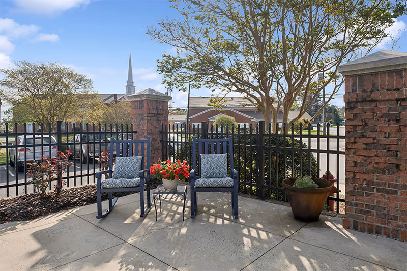 Outdoor patio area with two blue cushioned rocking chairs and a small table with a pot of red flowers between them. The patio is enclosed by a black metal fence and brick pillars. Trees and parked cars are visible in the background under a clear blue sky.