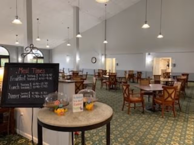 A spacious dining room with multiple wooden tables and chairs arranged neatly on a patterned green carpet. The room has high vaulted ceilings with hanging pendant lights and light gray walls. A small round table in the foreground displays a chalkboard sign listing meal times and some covered dishes.