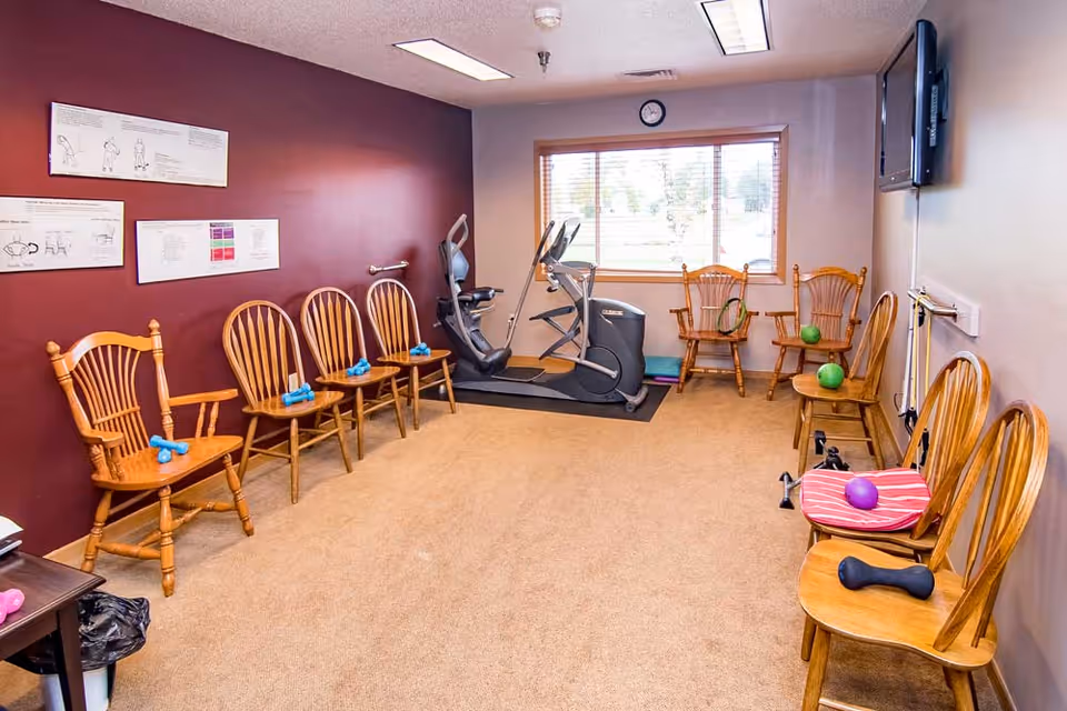 A small exercise room with wooden chairs arranged along the walls, each chair holding small hand weights. There is an elliptical machine near a window with blinds, and exercise instructions are posted on a maroon wall. A flat-screen TV is mounted on the opposite wall.