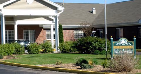 Exterior view of Woodmont Health Campus showing a brick building with a covered entrance, green bushes, and a lawn area. A sign in front reads 'Woodmont Health Campus'.