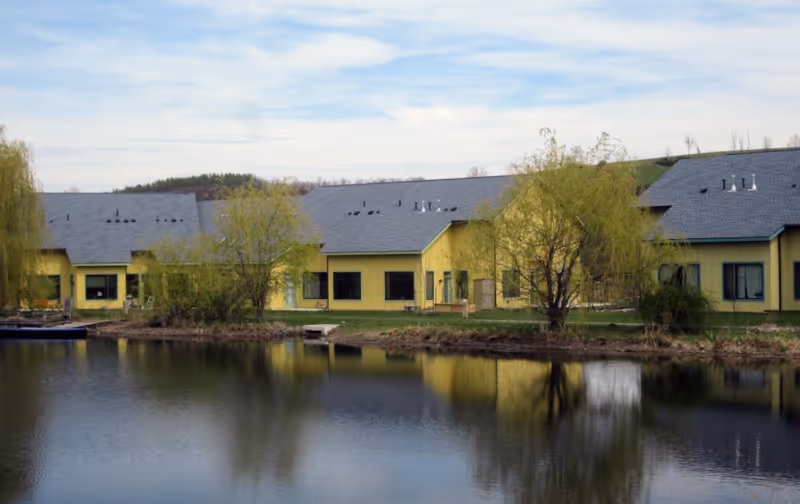 A calm pond reflecting a row of yellow buildings with gray roofs, surrounded by trees and greenery under a partly cloudy sky.
