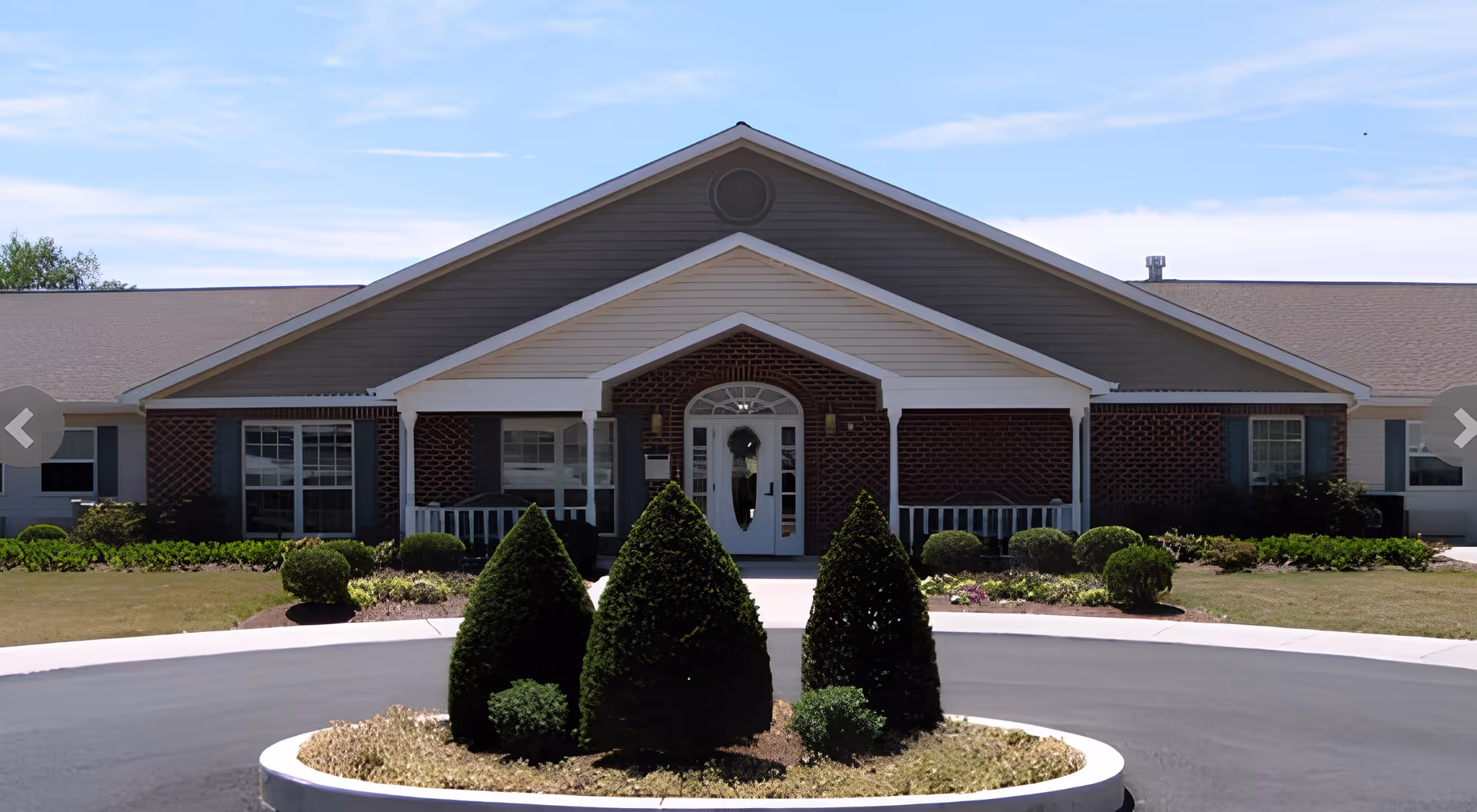 Front exterior view of Arden Courts - ProMedica Memory Care Community (Susquehanna) building with a circular driveway and neatly trimmed bushes in the center. The building features a peaked roof, brick and siding facade, and a white entrance door with sidelights.