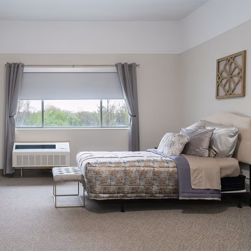 A neatly made bed with multiple pillows and a patterned comforter in a senior living bedroom. The room has a large window with gray curtains and a white roller shade, a wall-mounted air conditioning unit below the window, a small cushioned bench at the foot of the bed, and a decorative wooden wall art piece above the bed.